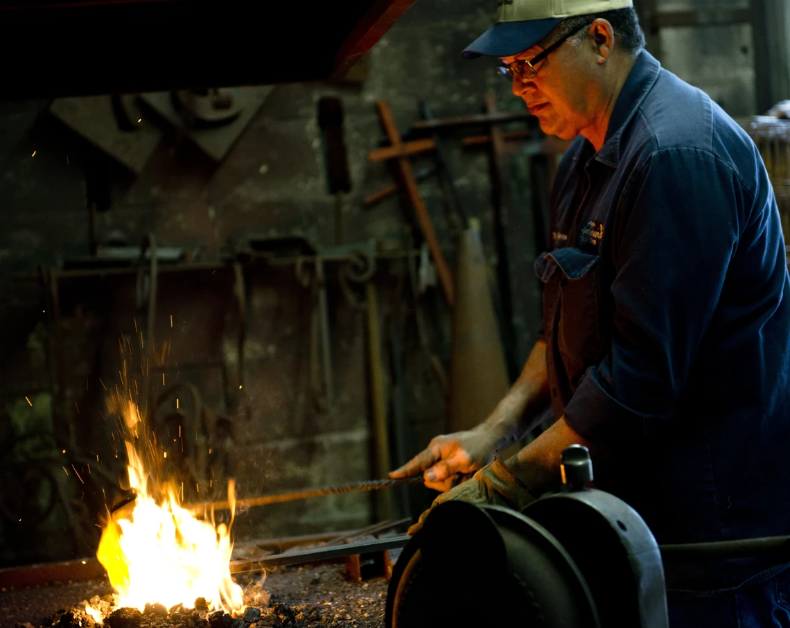 A man wearing a baseball cap and dark blue shirt heats a piece of metal in the fire of a coal-fired forge in his blacksmith’s shop.