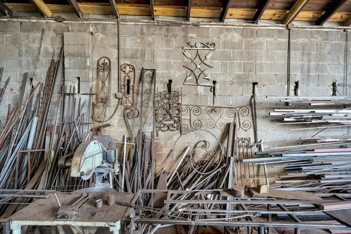 Numerous metal scrolls, pieces of ornamental ironwork, and long straight rods of metal fill the grey concrete wall of a blacksmith’s shop.