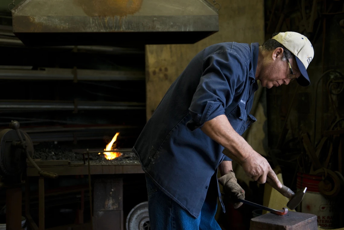 A man wearing a baseball cap and dark blue shirt works at an anvil beating a piece of metal with a hammer; his coal-fired forge with the flames of a small fire is behind him.