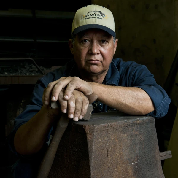 A man wearing a baseball cap that says Andrew’s Blacksmith Shop sits behind a dark metal anvil and holds a hammer in his hands.