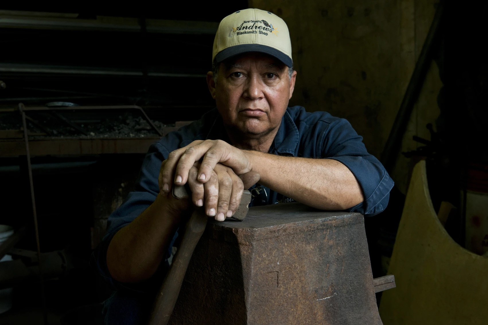 A man wearing a baseball cap that says Andrew’s Blacksmith Shop sits behind a dark metal anvil and holds a hammer in his hands.
