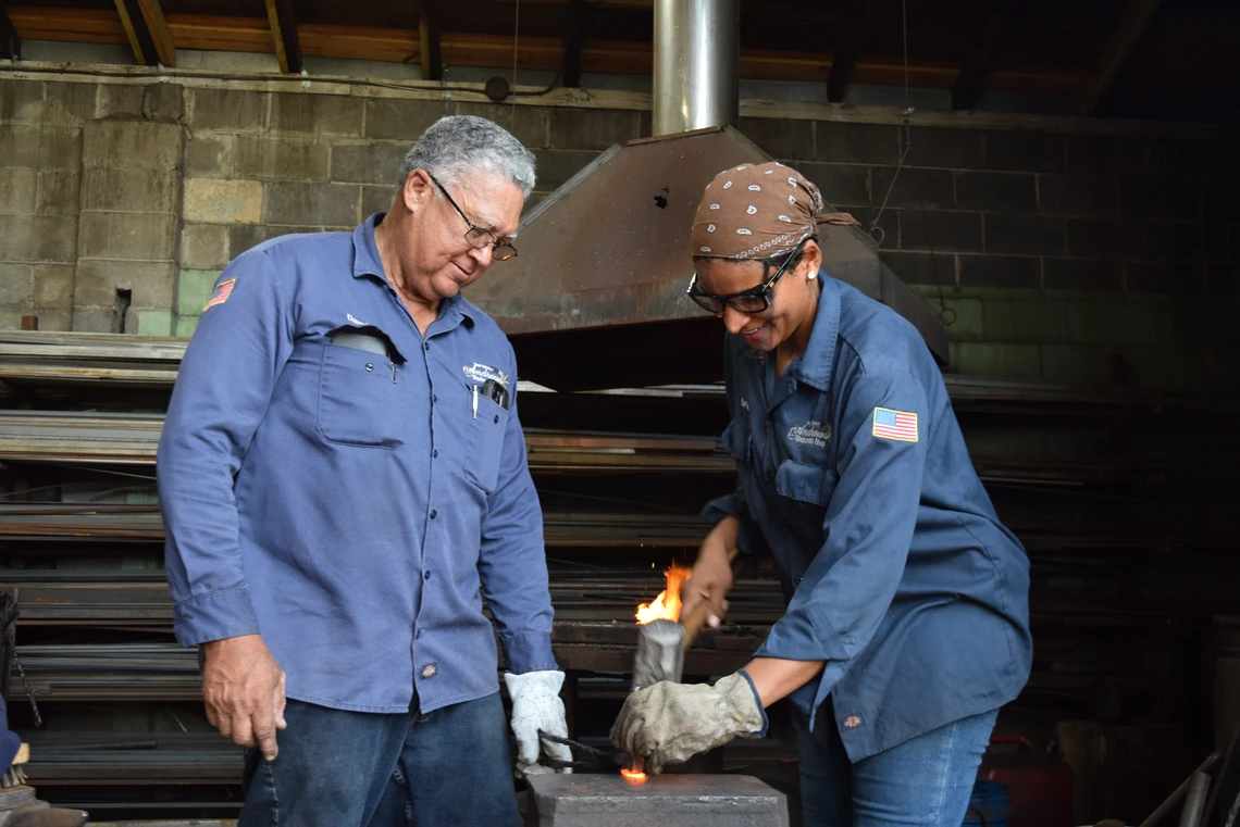 An older man and a young woman, both dressed in dark blue work clothes, work together at an anvil to shape a piece of metal with a hammer.