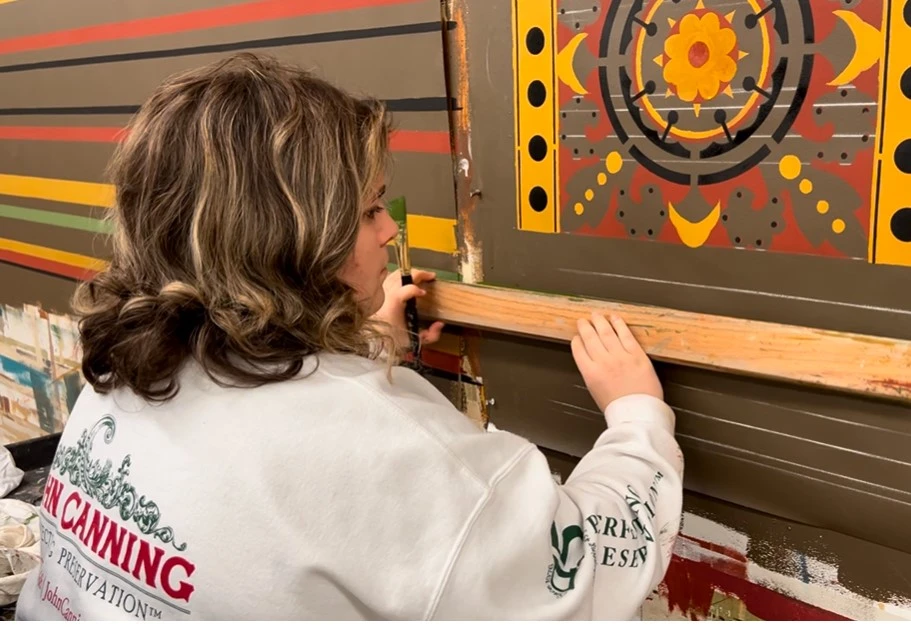 A young woman paints a green stripe on a wall underneath an orange, yellow, black, and tan geometric decorative motif.