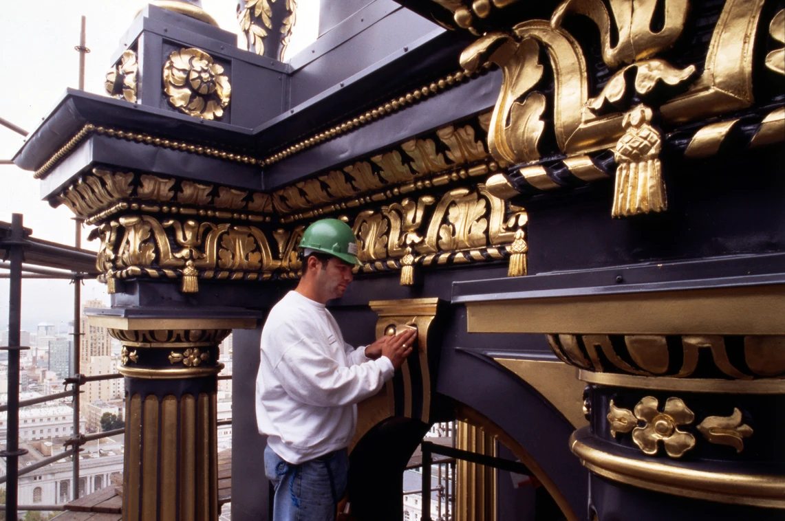 A man in a hard hat stands on scaffolding applying gold-leaf to ornamental elements on the exterior of a building.