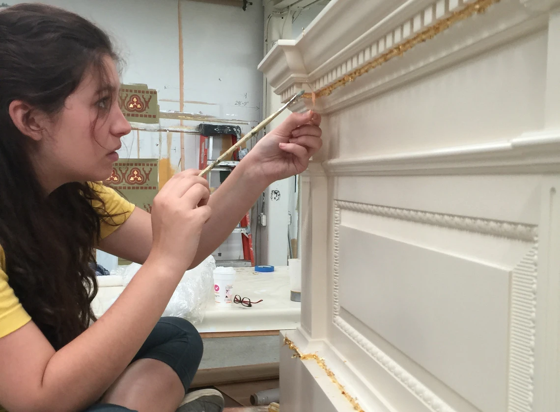 A young woman uses a small paint brush to apply gold leaf to a decorative molding.