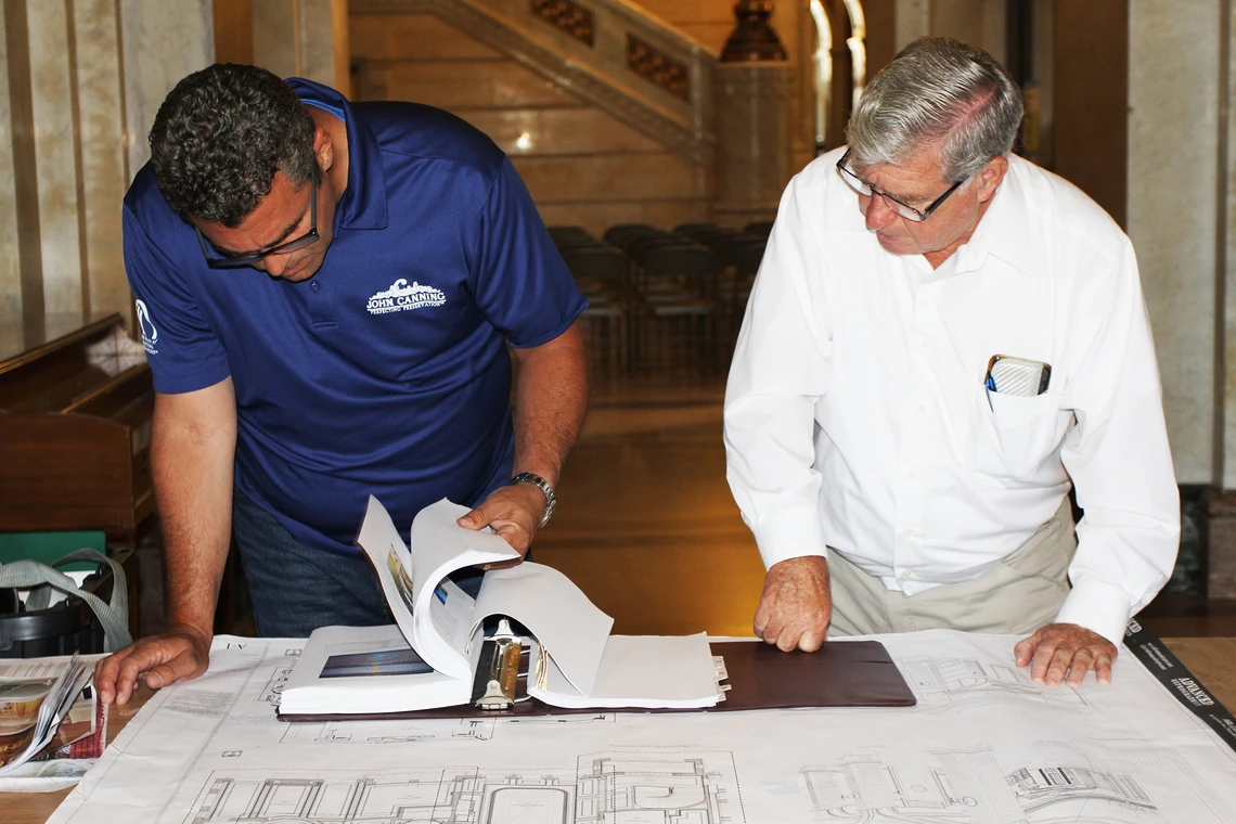 Two men lean over to look through the pages of thick binder of photographs and planning documents that is on a worktable topped with a large architectural drawing.