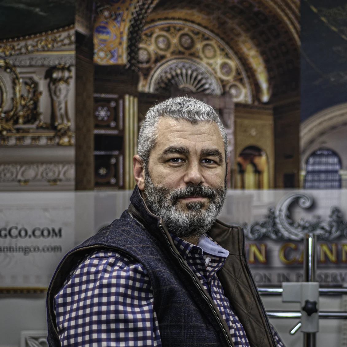 A man with grey hair and a beard poses in front of a display of decorative paintings.