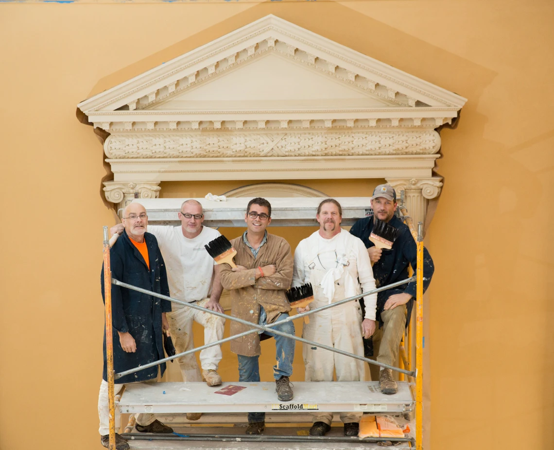 A group of five men holding paint brushes pose in scaffolding in front of an ornamental pediment in the interior of a building. 