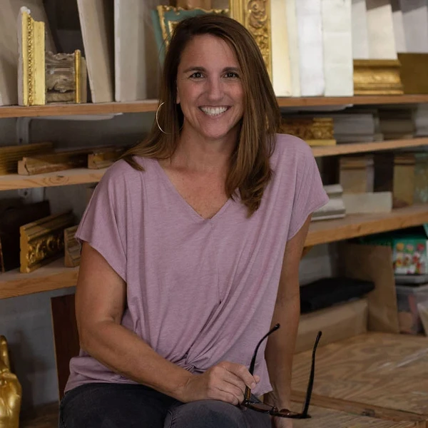 A woman in a light pink shirt sits in her studio in front of shelves filled with pieces of wood and decorative moldings.