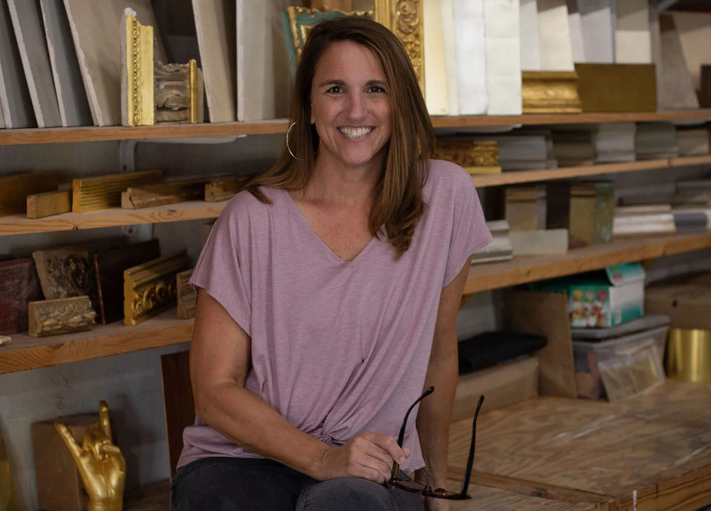 A woman in a light pink shirt sits in her studio in front of shelves filled with pieces of wood and decorative moldings.