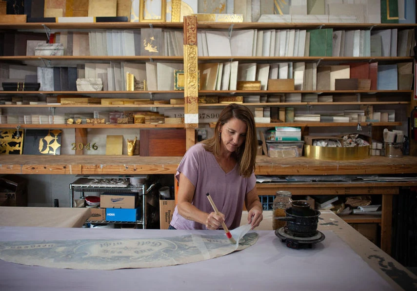 A woman works at a table in her studio to restore a blue floral decorative painting.
