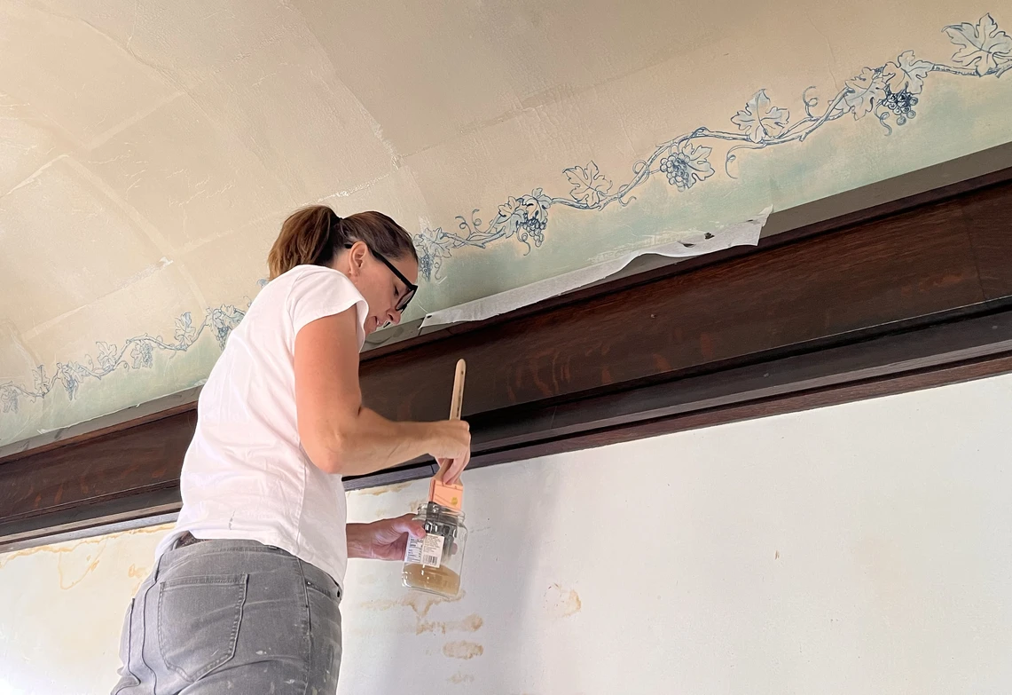 A woman uses a paint brush to restore a grape vine decorative painting running along the ceiling of a building.