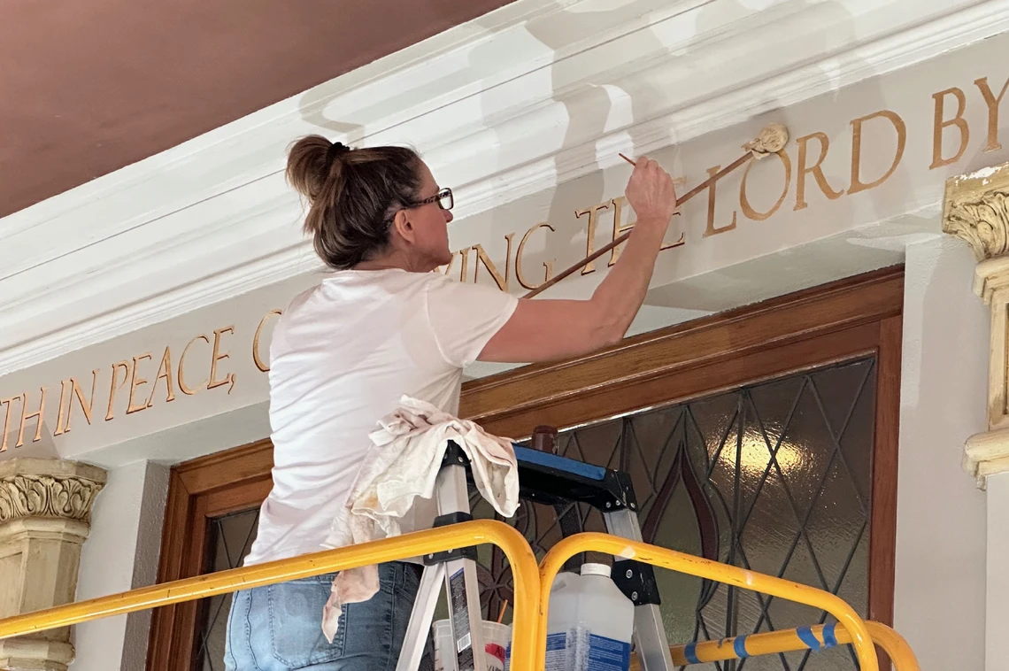 A woman stands on a scaffold painting gold letters for an inscription that runs along the top of an interior church wall.