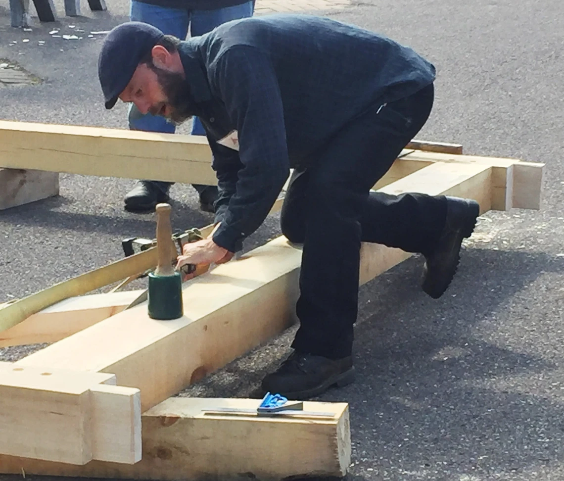 A man in a dark blue woolen cap and work clothes kneels to work with a mallet on a long, squared-off wooden beam.