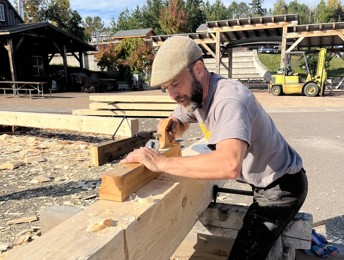 A man in a beige woolen cap and light grey t-shirt uses a wooden plane to smooth the surface of a large, long piece of timber positioned horizontally outside on blocks of wood; stacks of wooden beams are behind him.