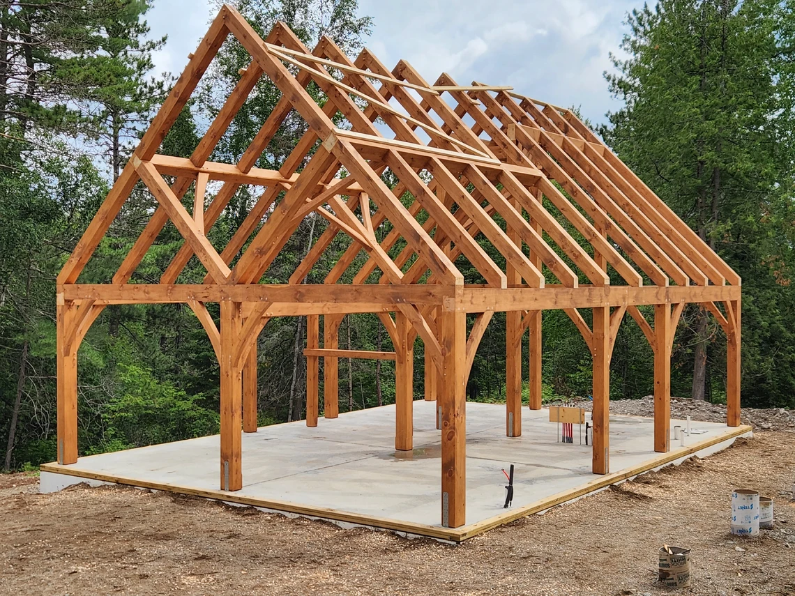 The skeleton-like timber-frame structure of a cabin features eleven large triangular wooden beams forming a roof and a series of vertical wooden beams for the walls.
