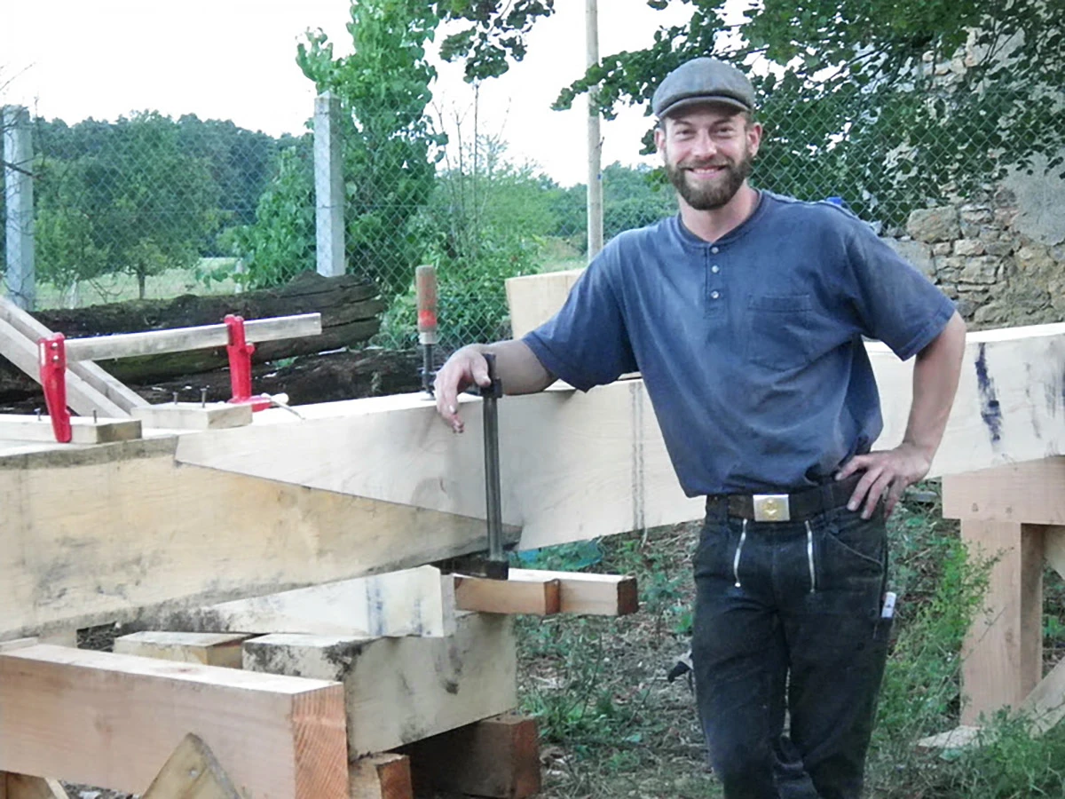 A man with a smile on his face poses outside in front of a large heavy wooden timber positioned horizontally on sturdy wooden sawhorses.