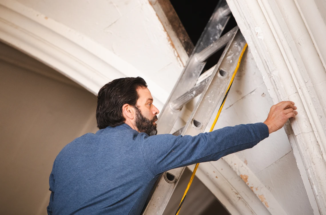 A man on a ladder reaches over to inspect the plaster moldings of an arch in the interior of a church. 