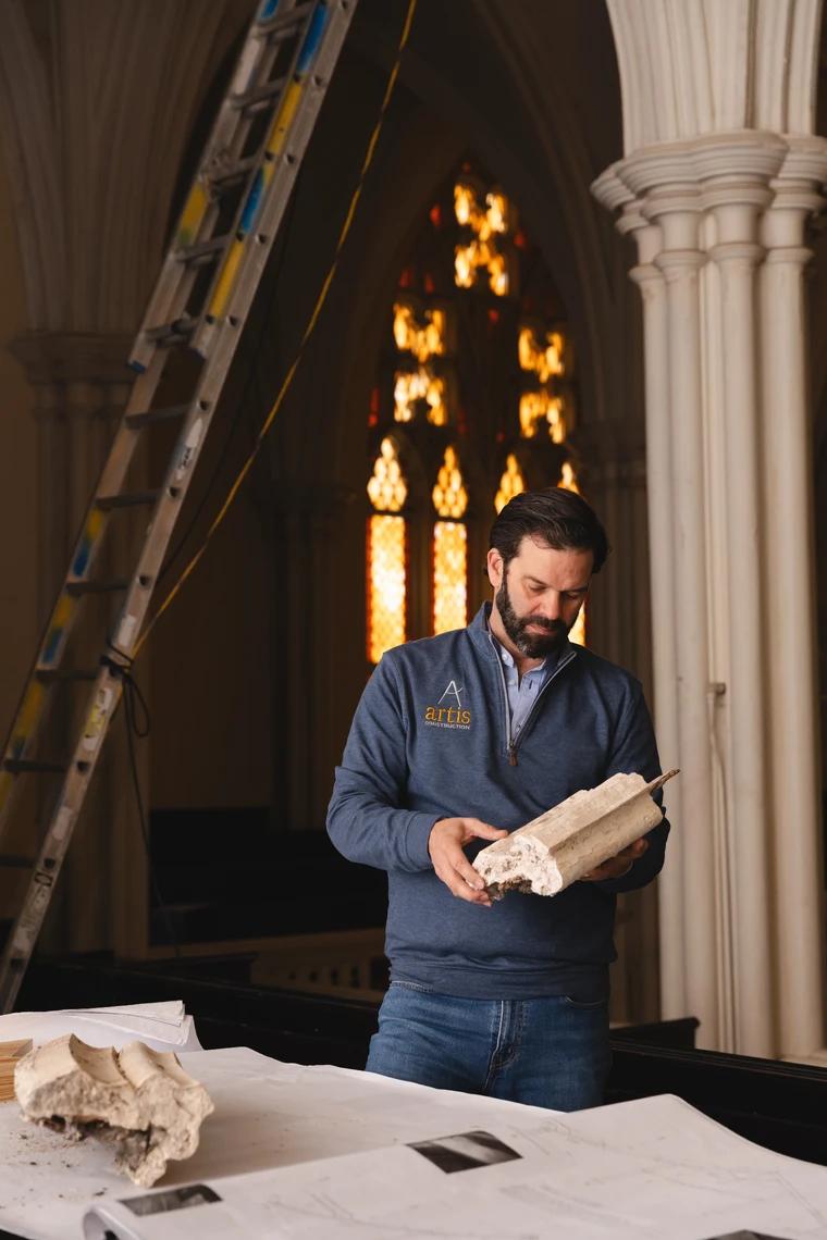 A man stands inside a church in front of a stained-glass window looking at a piece of damaged plaster molding.