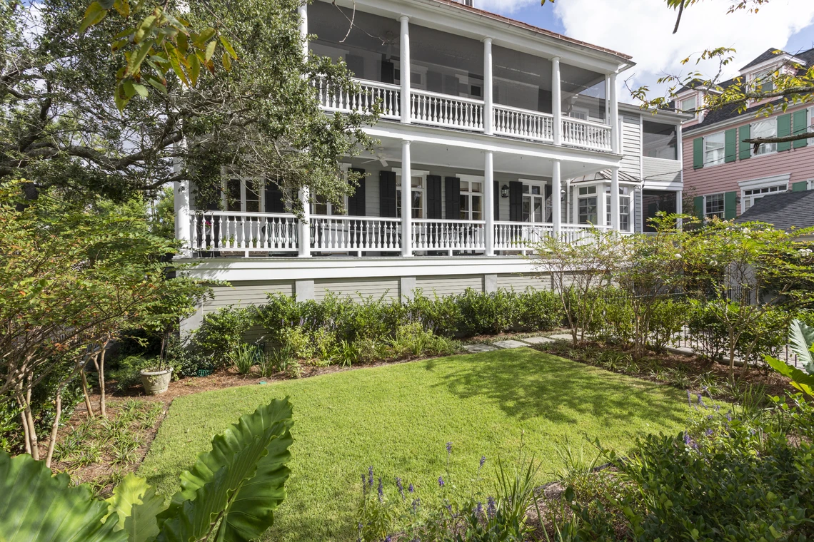 A big house with a large two-story porch running the length of the house stands in a lush green garden. 