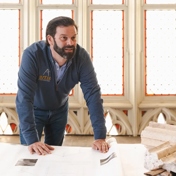 A young man leans over a table topped with architectural drawings and old pieces of ornamental plaster; church windows and stone arches are behind him.