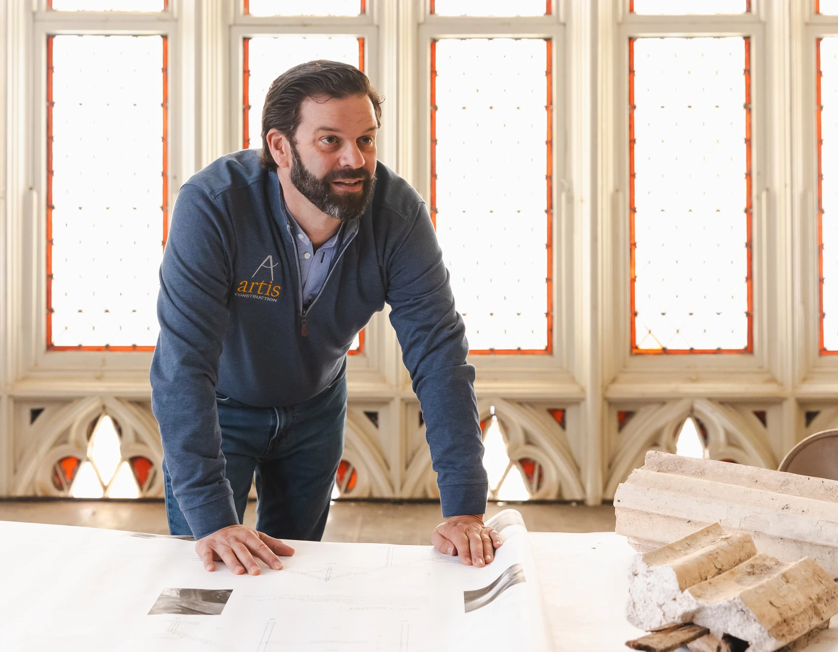 A young man leans over a table topped with architectural drawings and old pieces of ornamental plaster; church windows and stone arches are behind him.