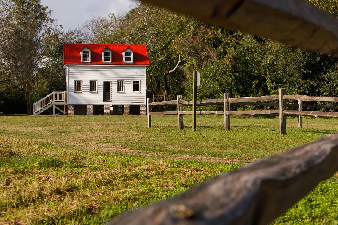 A white house with a bright red roof stands in a green field with trees behind it.