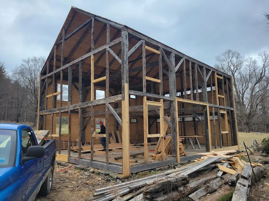 A large timber-framed skeleton of an old barn shows numerous original vertical and horizontal wooden beams mixed with new wooden beams used to replace damaged timber.
