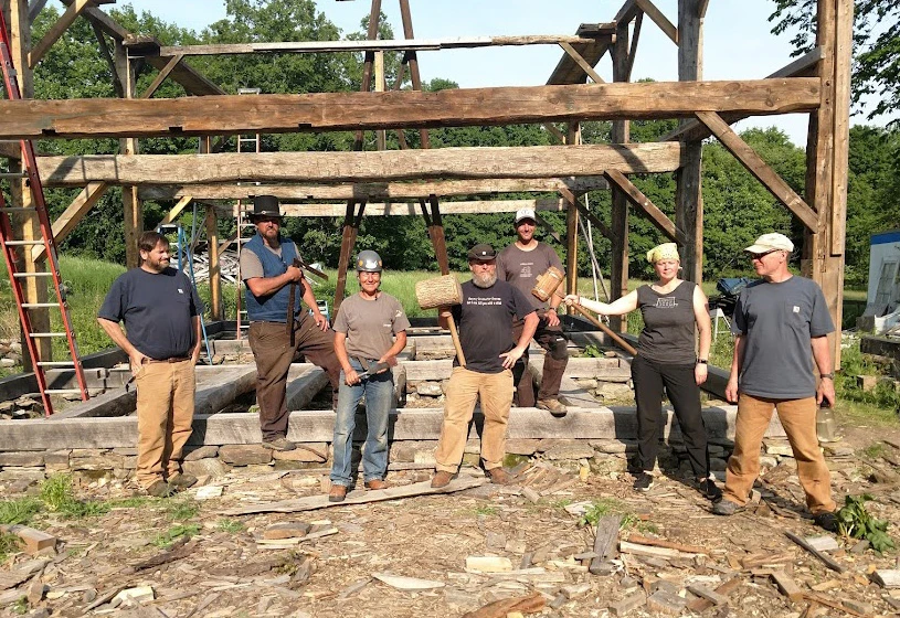 Seven people holding woodworking tools, including two very large wooden mallets, pose in front of the timber-framed skeleton of an old barn.