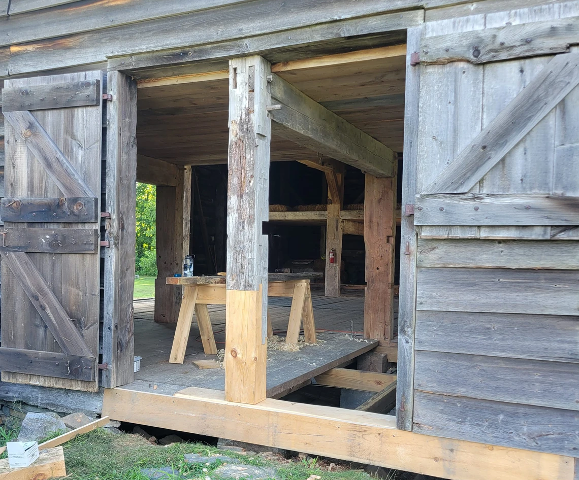 The opened wooden doors of an old historic barn reveal the interior of the barn with sawhorses and wooden beams inside.