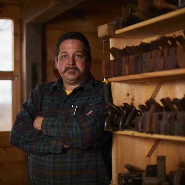 A man in a workshop poses in front of shelves filled with woodworking planes.