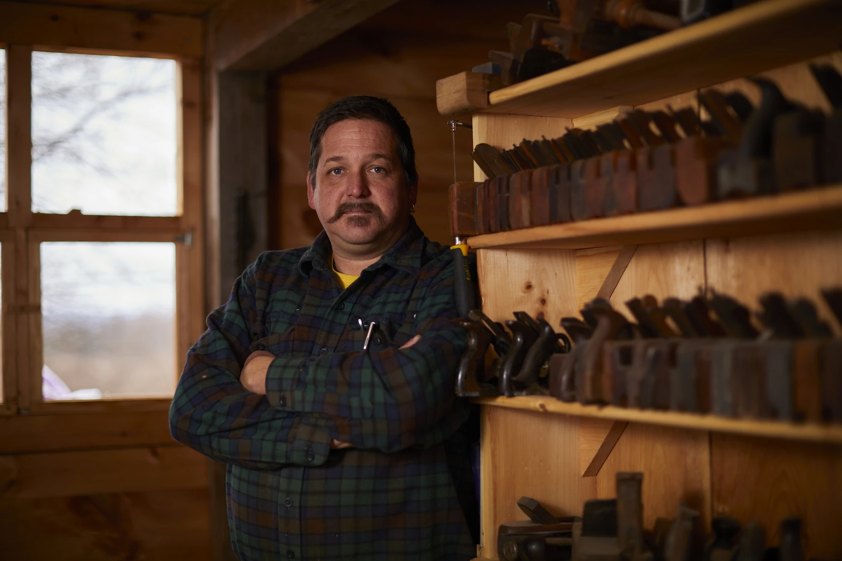 A man in a workshop poses in front of shelves filled with woodworking planes.