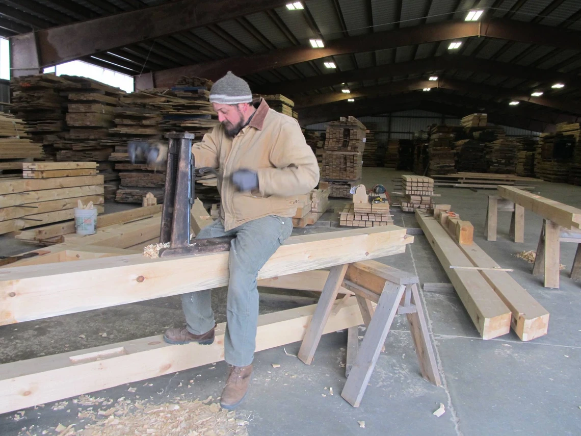 A man in large room filled with long, rectangular wooden beams is straddling a wooden beam and cutting a mortise joint in the wood with a metal hand-powered drill.