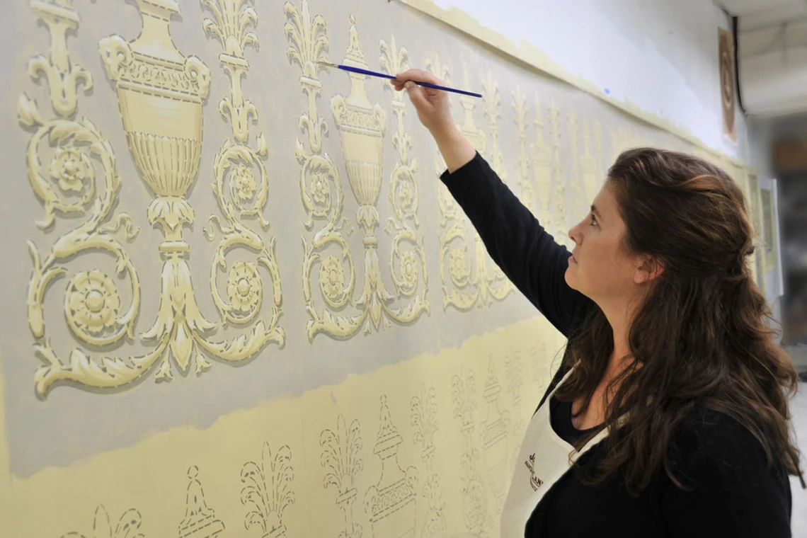 A young woman in an apron uses a small paintbrush to paint cream-yellow decorative scrolls and urns onto a canvas on the wall of a studio.