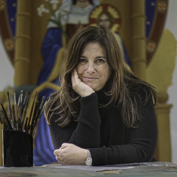 A woman in a studio sits at a table with a can of small paintbrushes next to her and a painting of a saint holding a branch of white lilies behind her on the wall.