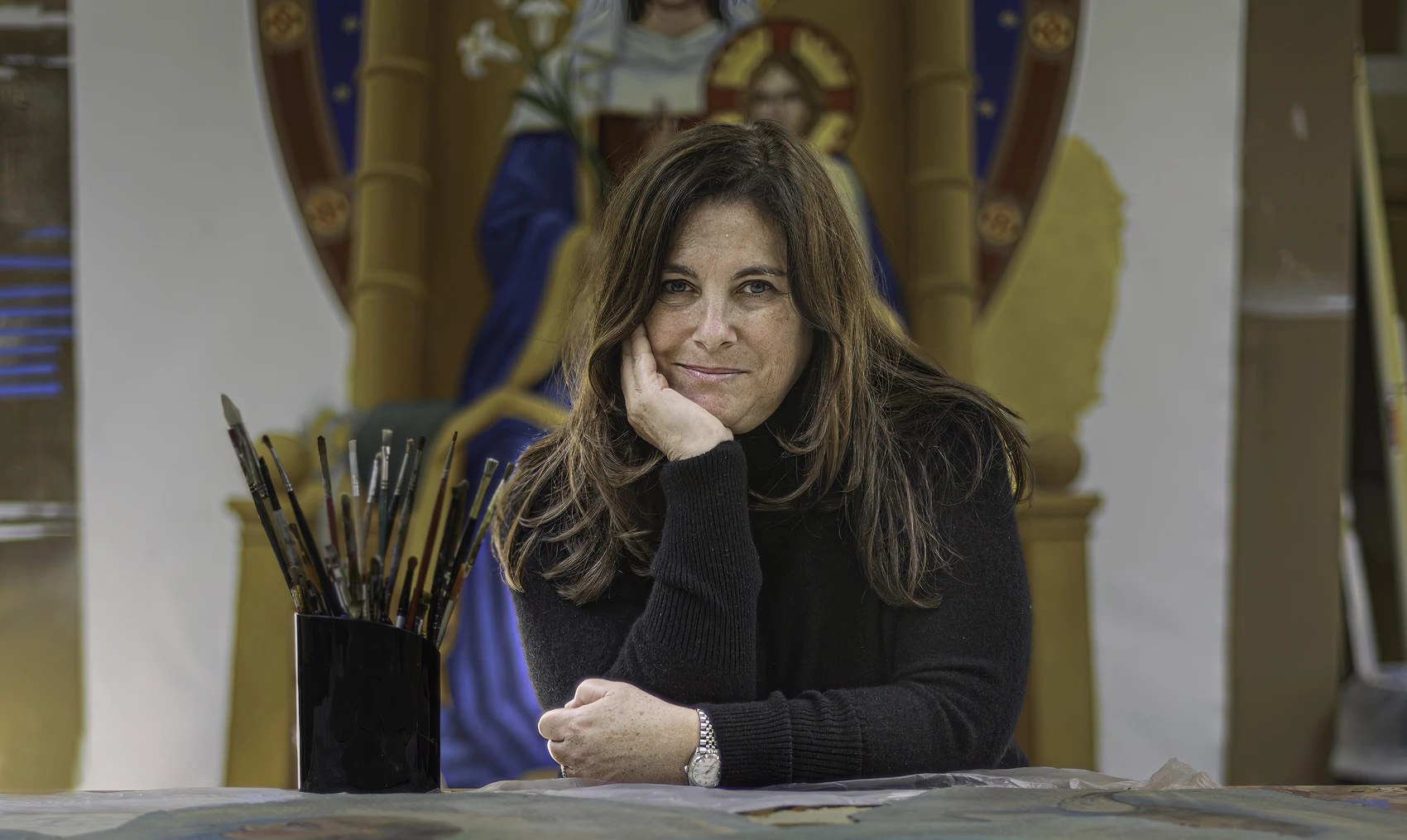 A woman in a studio sits at a table with a can of small paintbrushes next to her and a painting of a saint holding a branch of white lilies behind her on the wall.