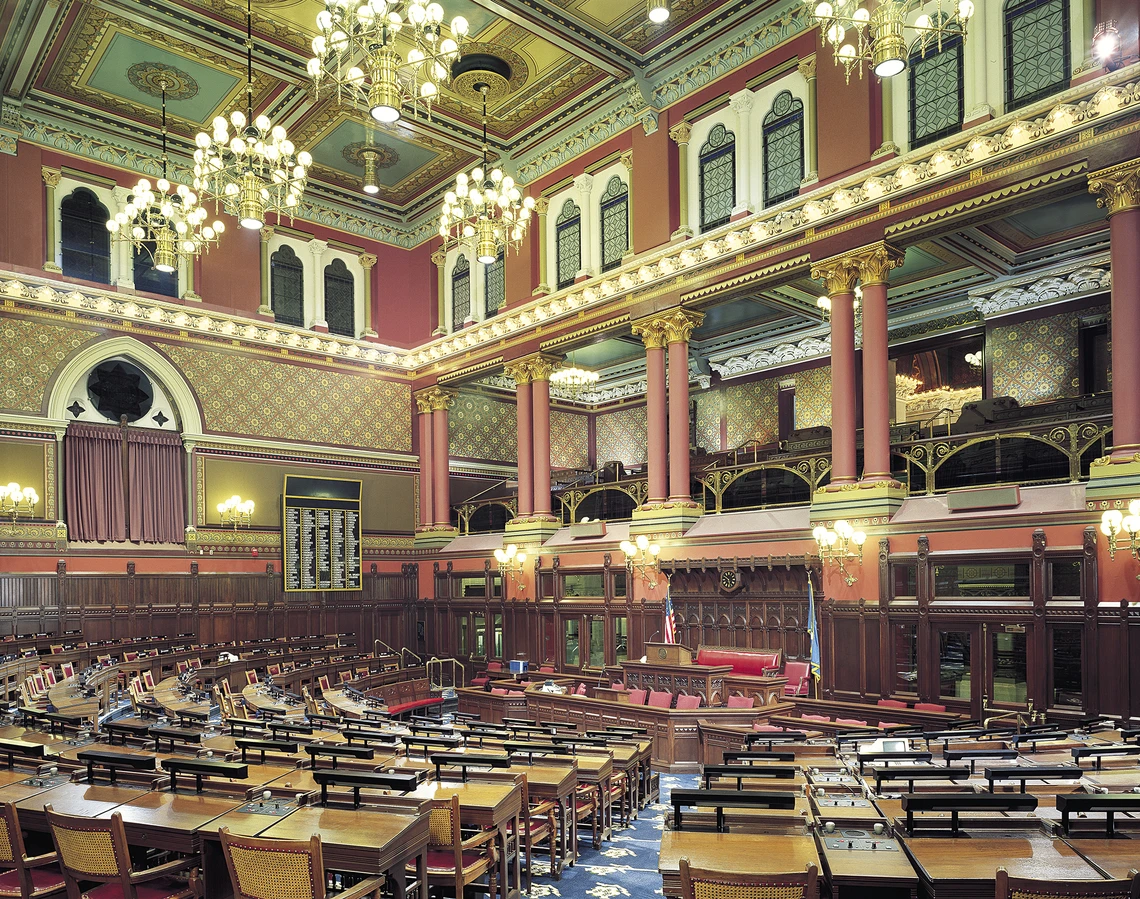 This image shows an elaboratively decorated room with rust-colored columns topped with gold Corinthian capitals, rust and gold-colored walls, arched windows with white trim, and chandeliers hanging from a green and gold ceiling. A multitude of wooden desks and chairs form a series of semi-circles on the floor of the room.