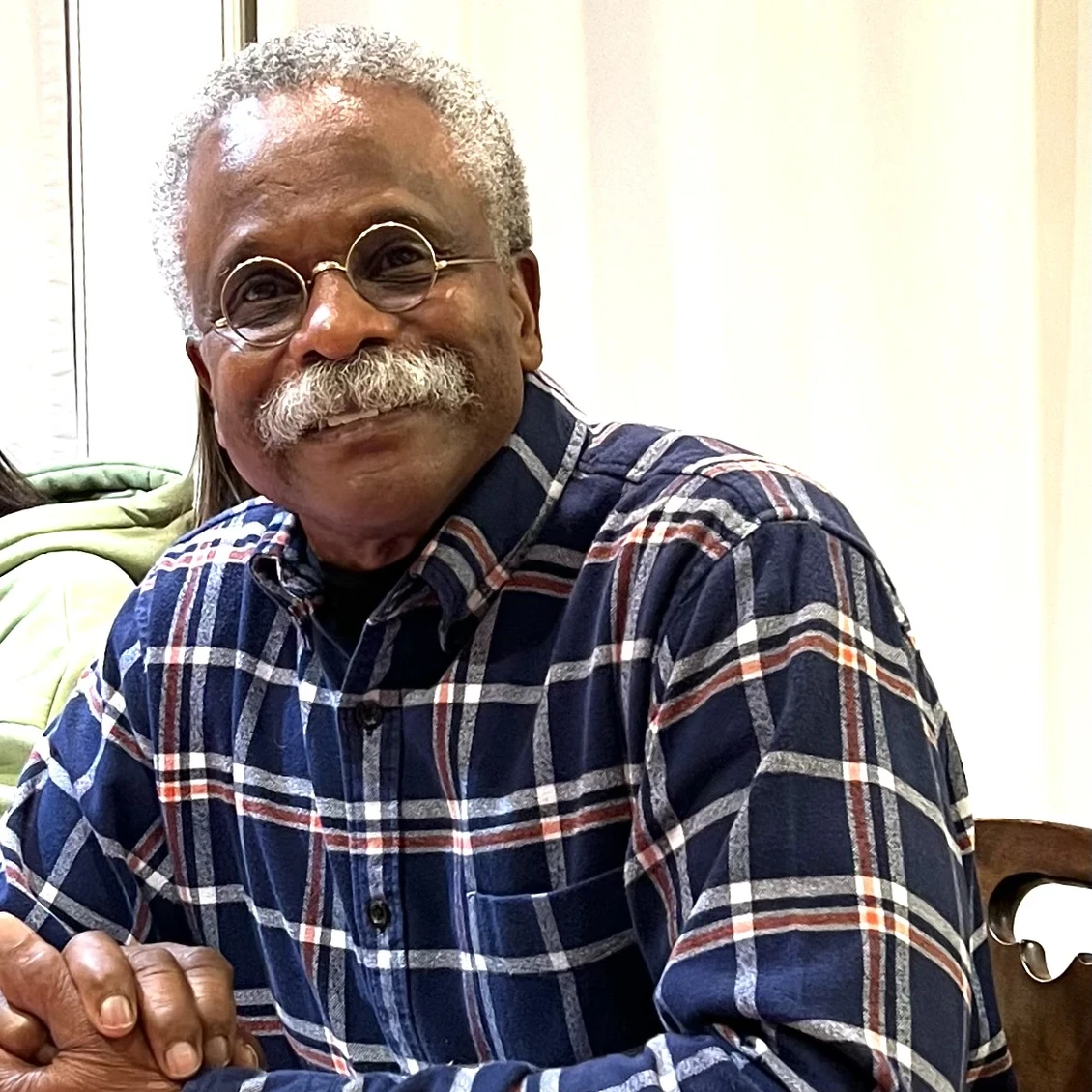 An older man with grey hair, a grey mustache, and round wire-rimmed glasses smiles warmly as he sits at a desk.