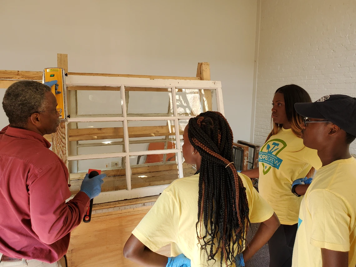 In a classroom setting, an older man in a red shirt and khaki pants shows three college students in yellow t-shirts how to restore an old window that has thin wooden strips separating nine small panes of glass; the window is mounted on wooden frame on a worktable.