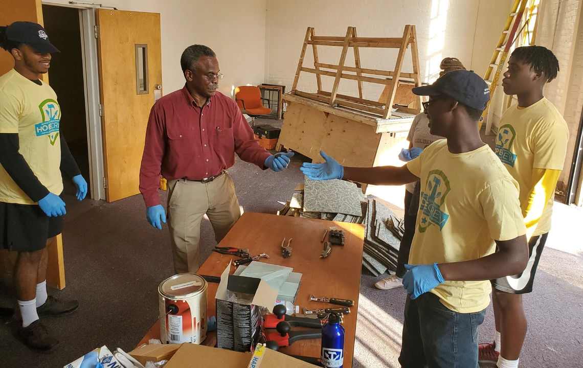 In a classroom setting, an older man in a red shirt and khaki pants shows a woodworking tool to a group of three young college students in yellow t-shirts gathered around a worktable topped with a bucket of paint and several tools.