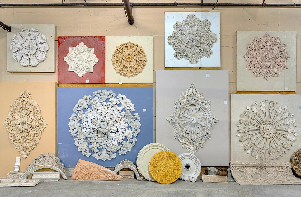 Elaborate white plaster ceiling medallions mounted on square wooden boards hang on the wall of a plastering shop.