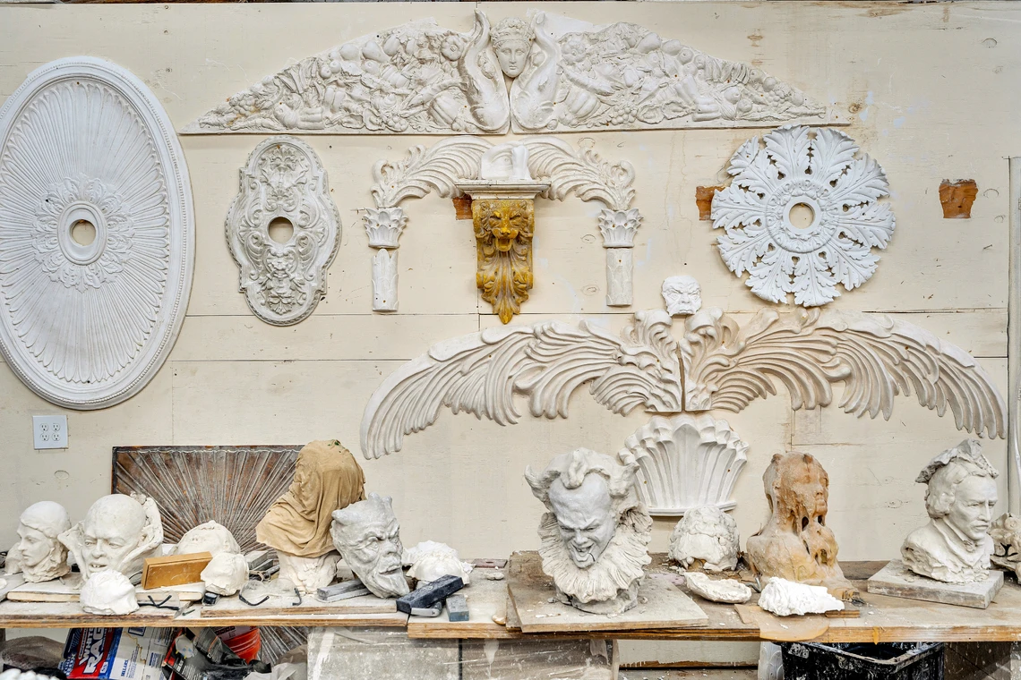 Numerous intricate white plaster ornamental pieces hang on the wall of a plastering shop; a wooden shelf lined with various plaster heads and busts runs along the bottom third of the wall.