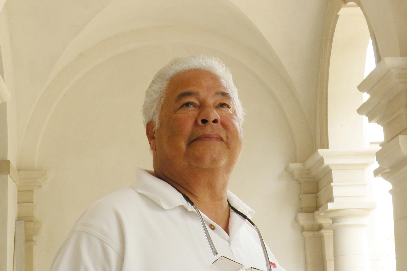 An older man with white hair and a white shirt stands under an arched ceiling made of smooth white plaster.
