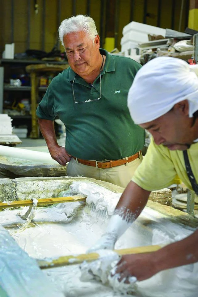 An older man with white hair watches a younger man work with his hands in wet white plaster as he tries to make a round plaster molding. 