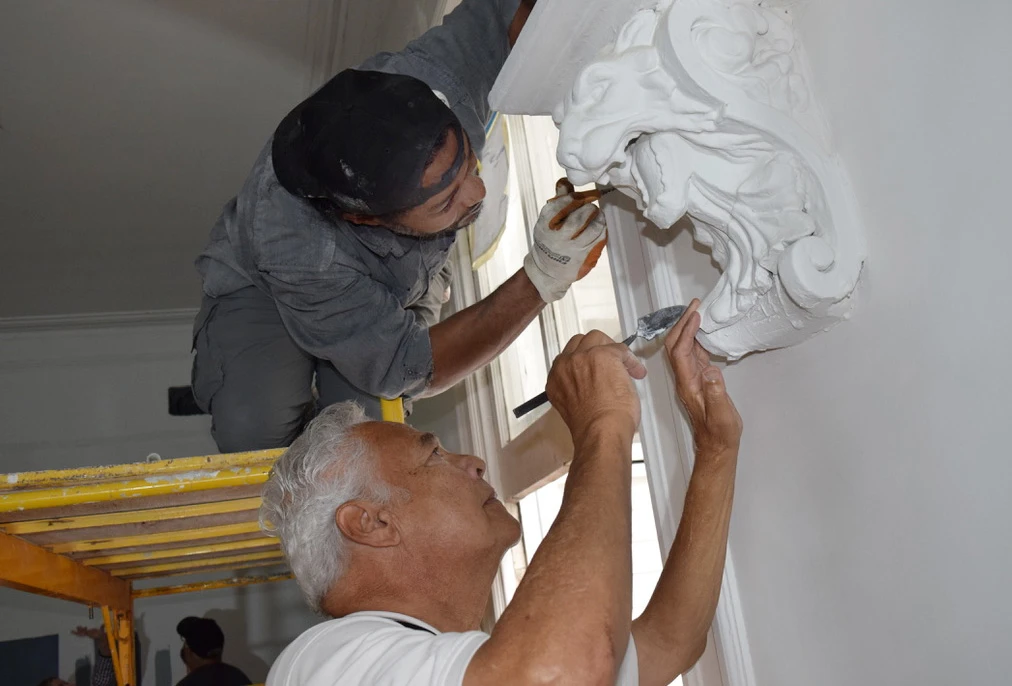A young man kneeling on a scaffold and an older man standing on the floor use small metal tools to put the finishing touches on a white plaster ornamental corbel of a lion located near the top of an interior plaster wall.