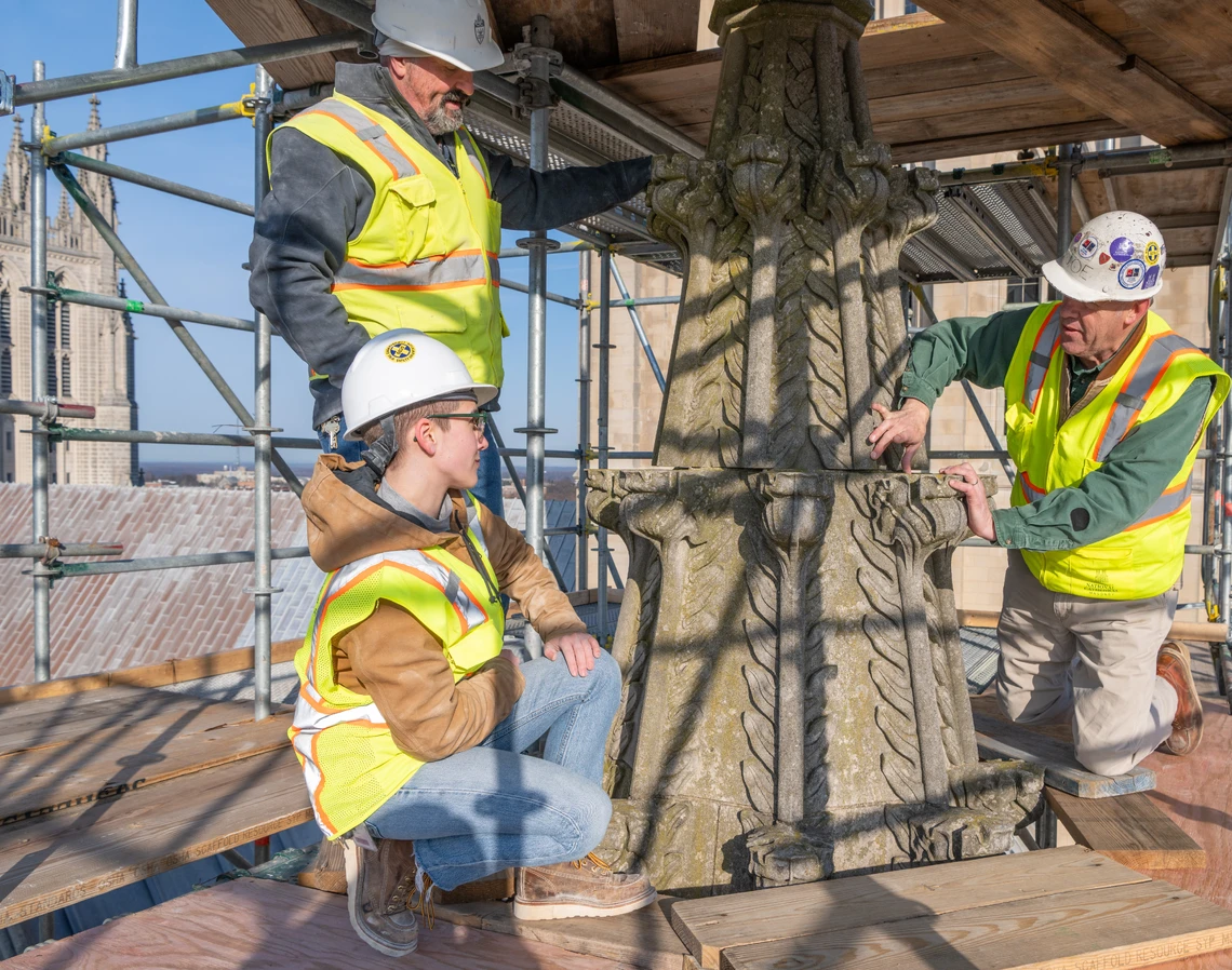 Atop a scaffold platform at a cathedral, a man in a white hard hat and yellow safety vest shows a young woman and a man what needs to be repaired on a large, damaged Gothic-style pinnacle stone.