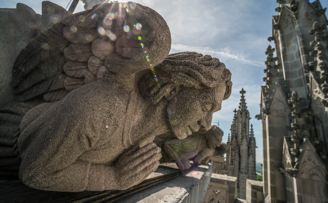 Hand-carved limestone angels with feathered wings and praying hands lie along the top of a stone balustrade awaiting reinstallation on a cathedral tower; the spires of two tall Gothic-style pinnacles are visible in the near distance. 