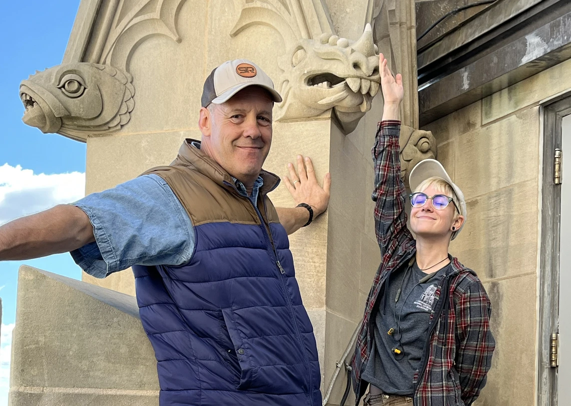 Atop a Gothic-style cathedral, a man smiles broadly as a young woman standing next to him proudly points to a gargoyle-like stone carving above her; a decorative carving of a fish is to the left of the man’s head.