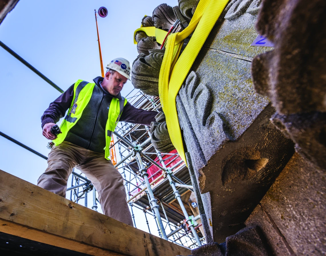 A man in a white hard hat and yellow safety vest stands on scaffolding while guiding a large elaborately carved Gothic-style limestone pinnacle rigged with yellow hoisting straps down to the ground.