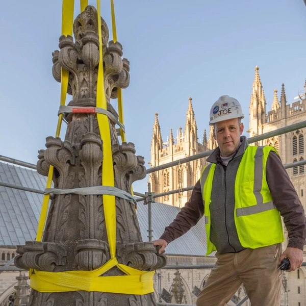 A man in a white hard hat and yellow safety vest stands on scaffolding next to a large elaborately carved Gothic-style limestone pinnacle rigged with yellow hoisting straps; the tall twin towers of a cathedral rise in the blue sky behind him.